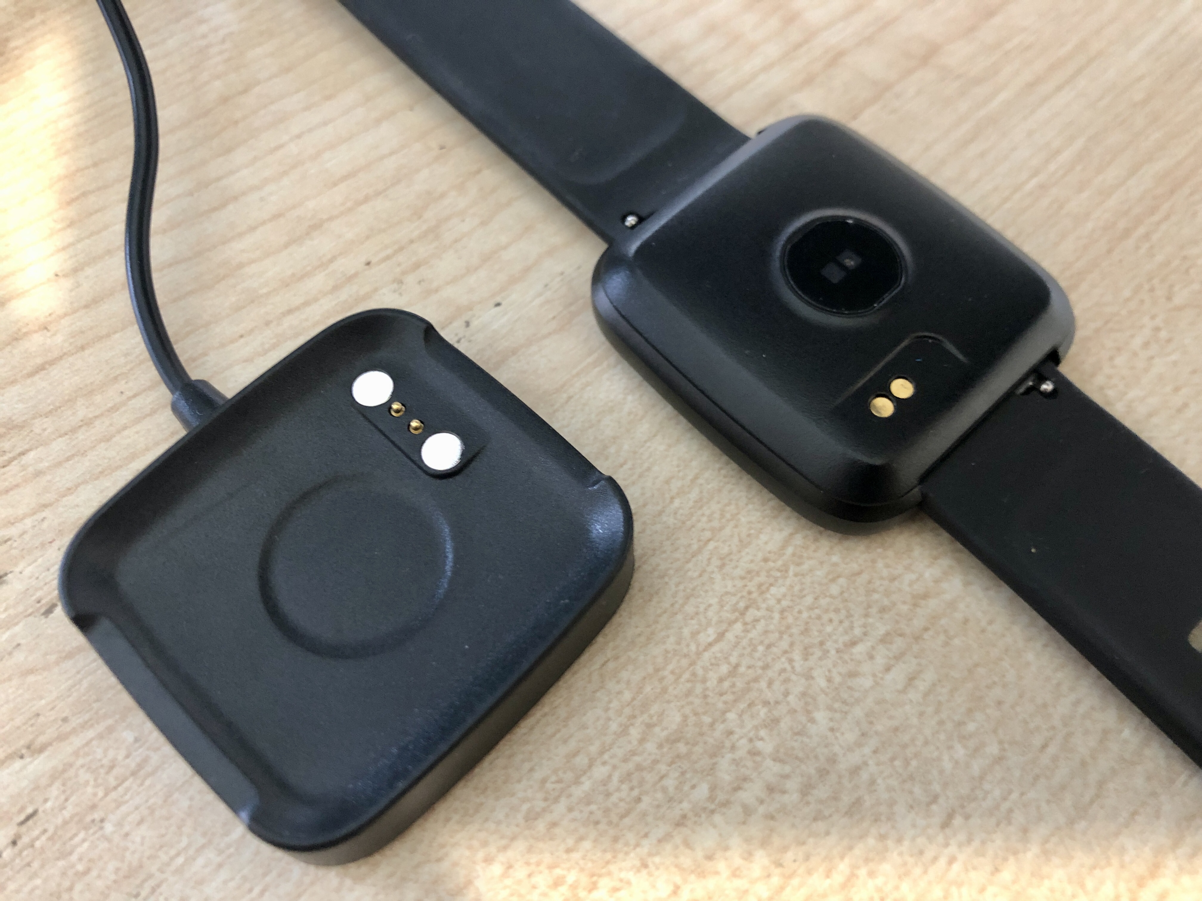 Photo of PineTime watch next to the small black square charging pad, on a wooden desk