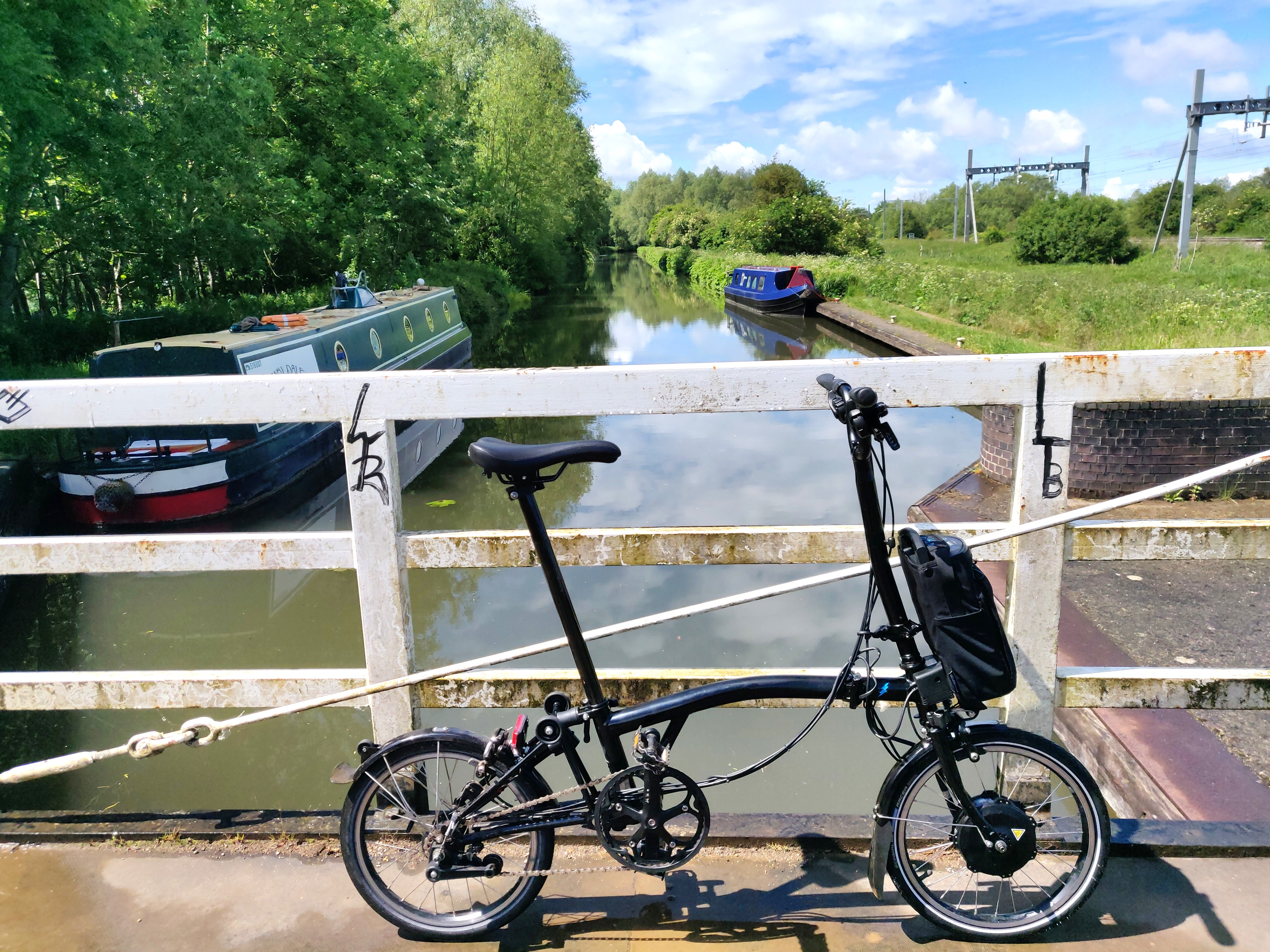 Photo of a black Brompton folding bike on a bridge over a canal. There is a green narrowboat moored to the left, and, in the distance, a blue narrowboat moored to the right. There is plenty of greenery on both sides of the canal. The water is a murky grey/green