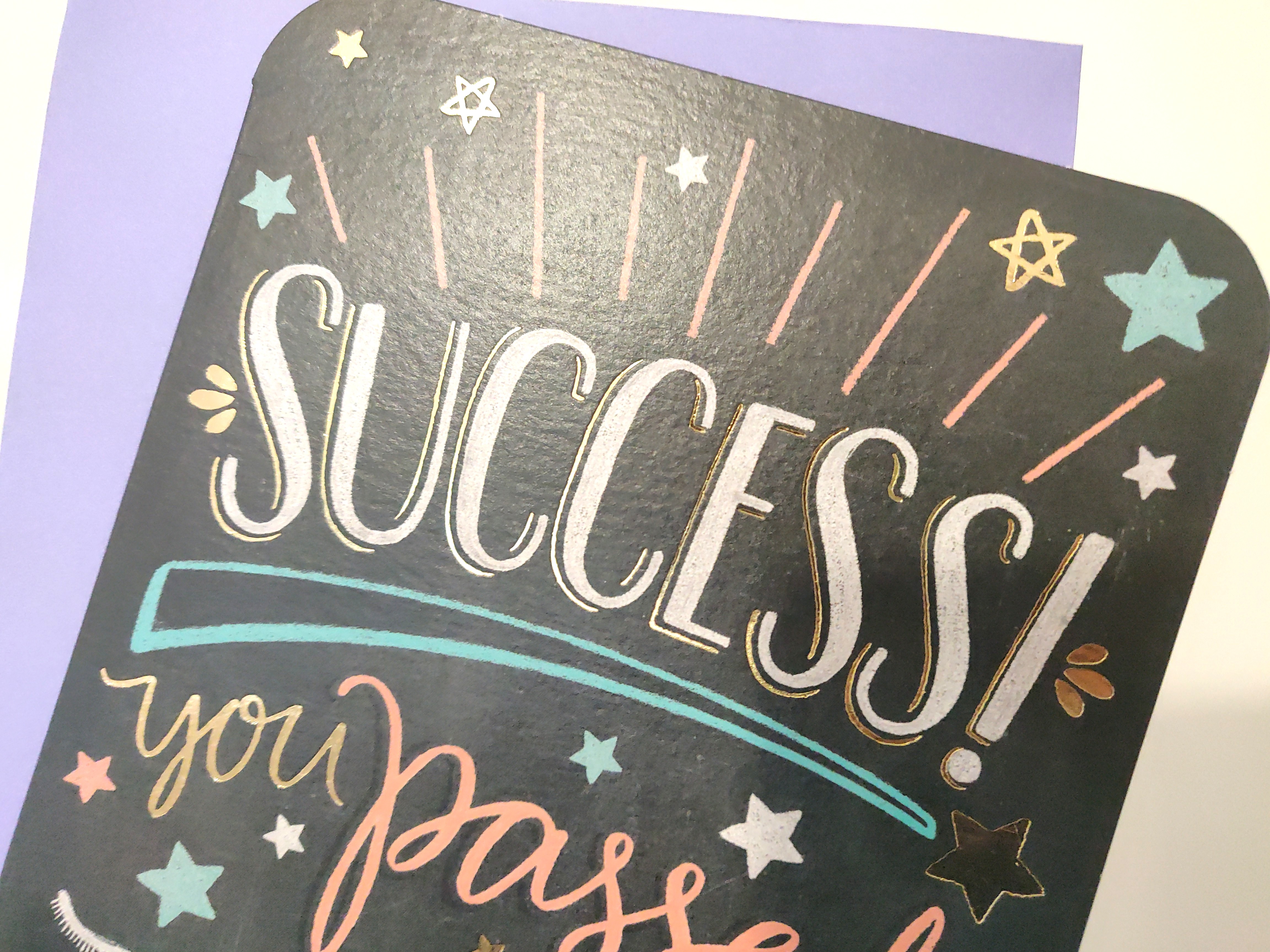 Bad photo of a card saying &ldquo;Success! You passed!&rdquo; in an exciting font, on a black background. There is a purple envelope in the background.