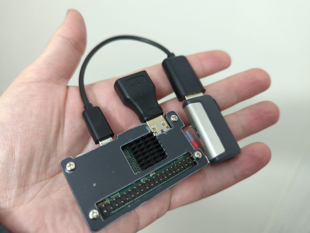 Photo of a Raspberry Pi Zero W, in the palm of my hand. There is a micro HDMI to HDMI adapter, and a micro USB to USB adapter with a USB memory stick. The Pi is a black plastic case, with a visible black heatsink
