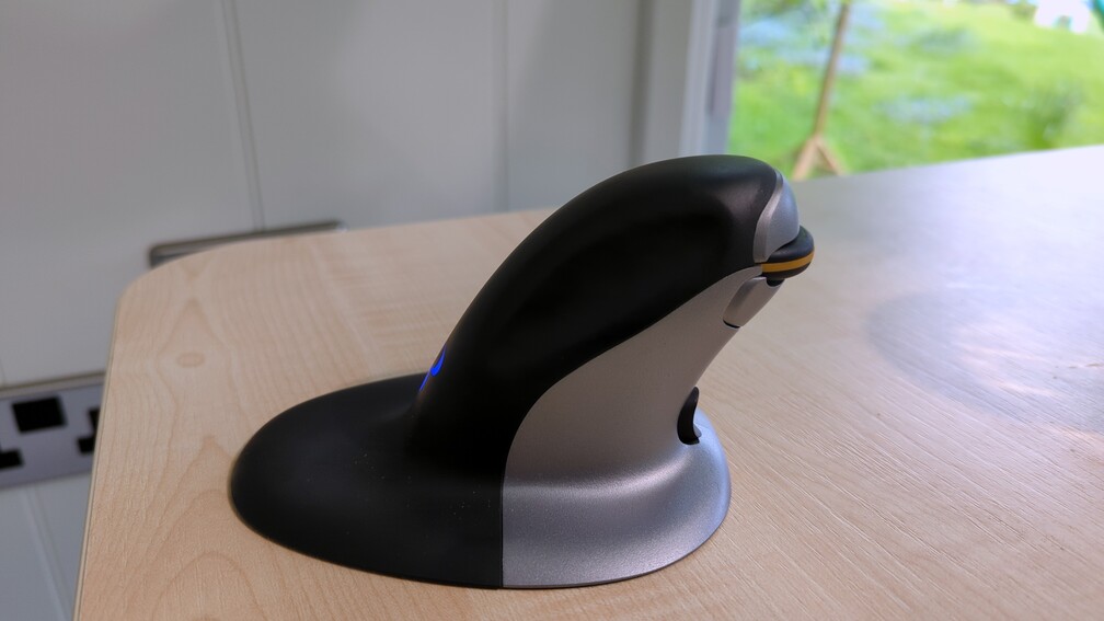 Photo of the black-and-silver tall mouse, with a large flared base, and a horizontal scroll wheel at index finger height, resting on a desk made of light coloured wood